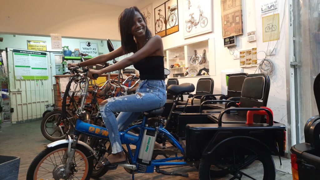 אישה יושבת על אופניים חשמליים בחנות, מחייכת עם סביבה של אביזרי אופניים וציוד לתחזוקה.